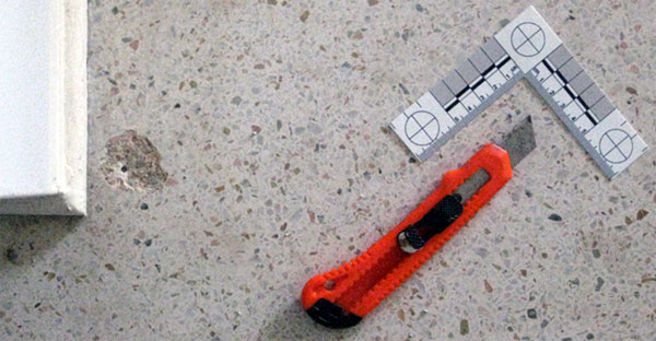 A view from above of an orange box cutter and 2-arm forensic scale on the floor near a wall corner